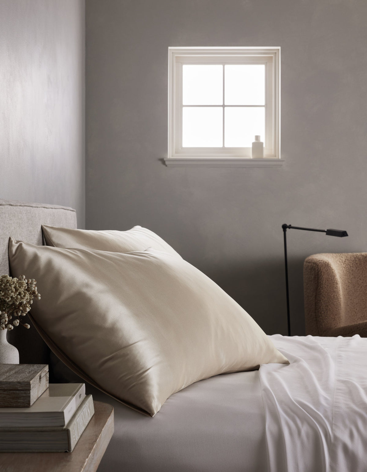 A minimalist bedroom features a neatly made bed with Cozy Earth's Silk Pillowcases, large cream-colored pillows, a nightstand holding books and dried flowers, soft light from a small window, and a beige chair in the background.|Color:Oat