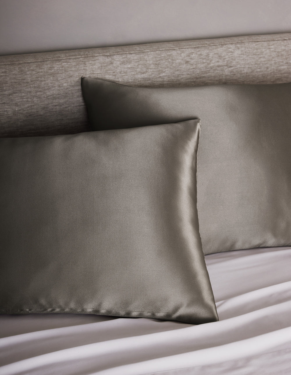 Two Cozy Earth Silk Pillowcases are placed on a bed with a light gray upholstered headboard and white sheets, adding a modern and elegant touch.|Color:Charcoal