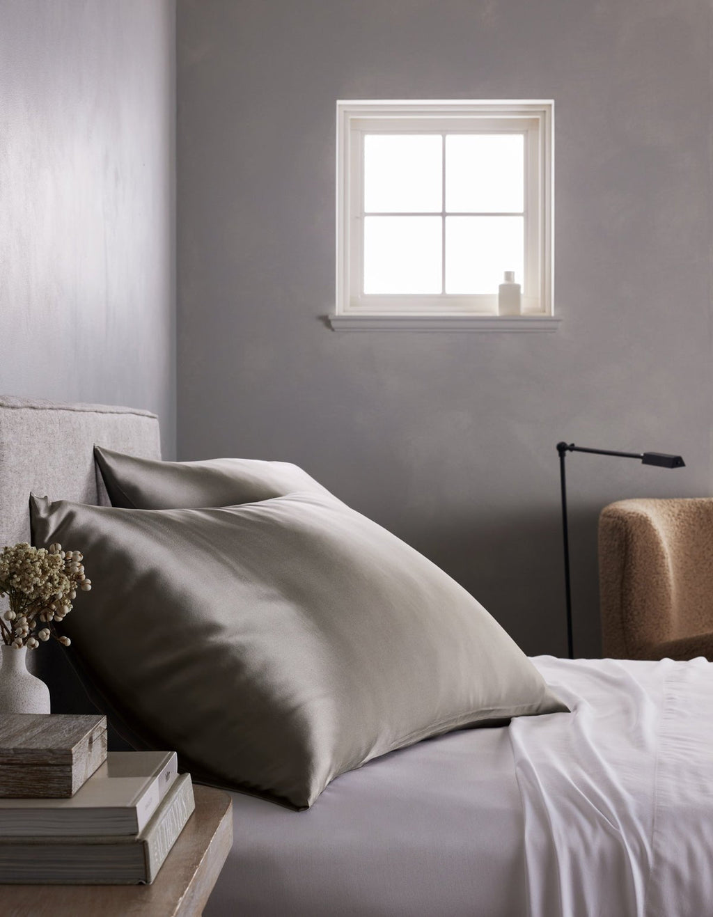 A minimalist gray bedroom features a neatly made bed with large Cozy Earth Silk Pillowcases in gray and white sheets, set beside a small stack of books, dried flowers, a beige chair, and a window that lets in natural light.|Color:Charcoal