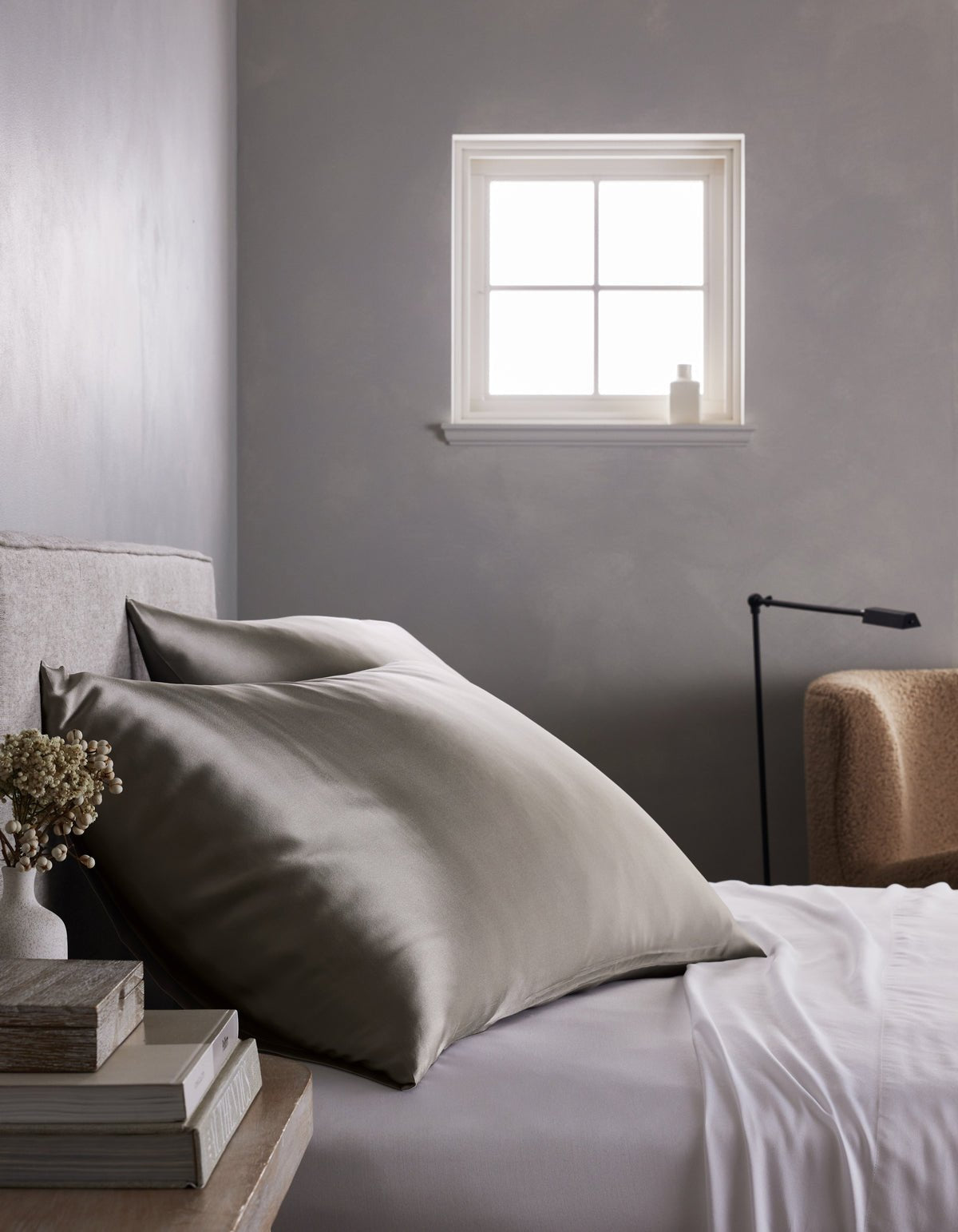 A minimalist gray bedroom features a neatly made bed with large Cozy Earth Silk Pillowcases in gray and white sheets, set beside a small stack of books, dried flowers, a beige chair, and a window that lets in natural light.|Color:Charcoal