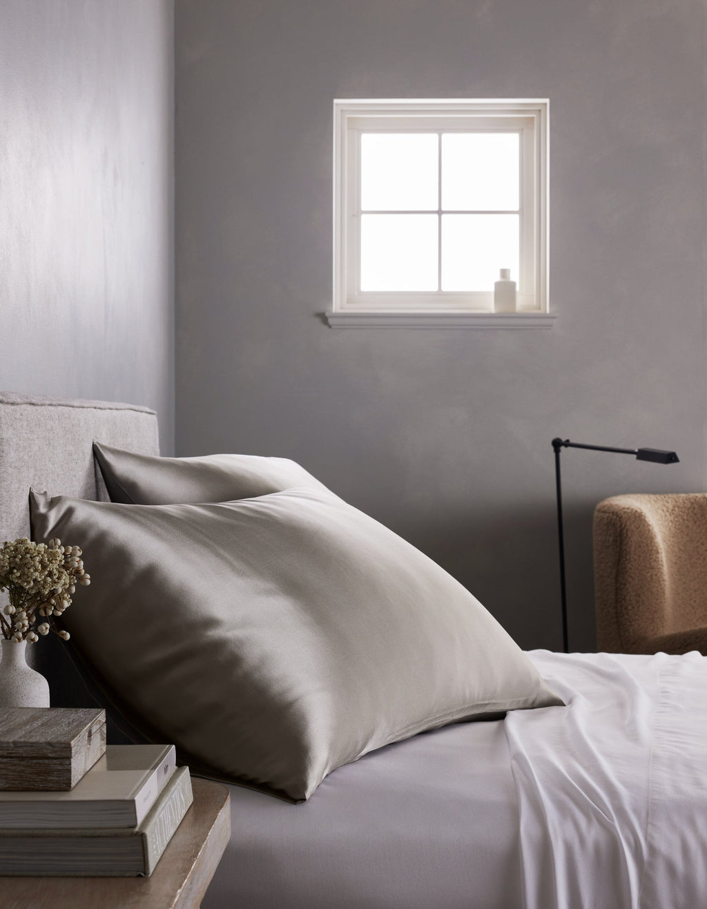 A neatly made bed features large, smooth Cozy Earth Silk Pillowcases in silver-gray and crisp white sheets beside a small table with books and flowers. A soft chair, lamp, and window on a gray wall complete the minimalist bedroom.|Color:Dove Grey