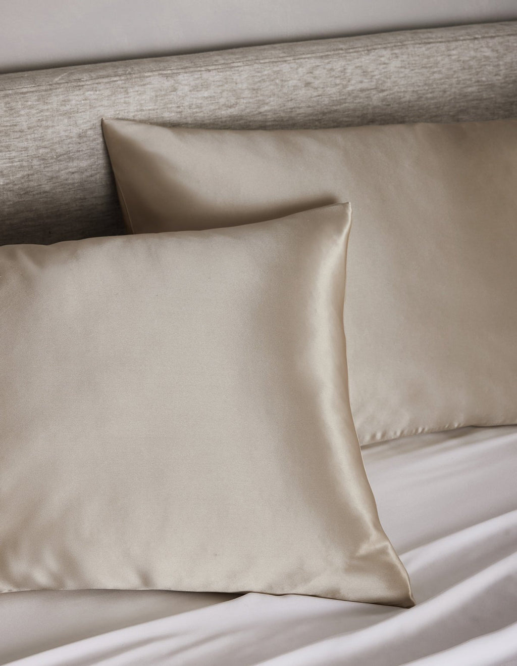 Two Cozy Earth Silk Pillowcases in light beige are displayed on a bed with a matching headboard and white sheets, creating a soft and elegant bedding arrangement.|Color:Driftwood