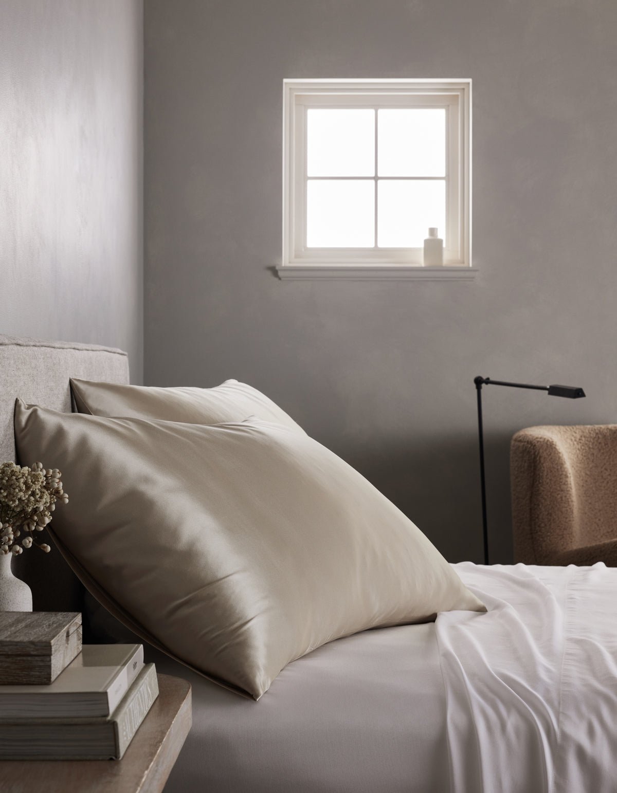 A neatly made bed features large cream Cozy Earth Silk Pillowcases and a white sheet beside a small window. A stack of books and dried flowers sit on the bedside table, with a beige armchair and black lamp in the background.|Color:Driftwood