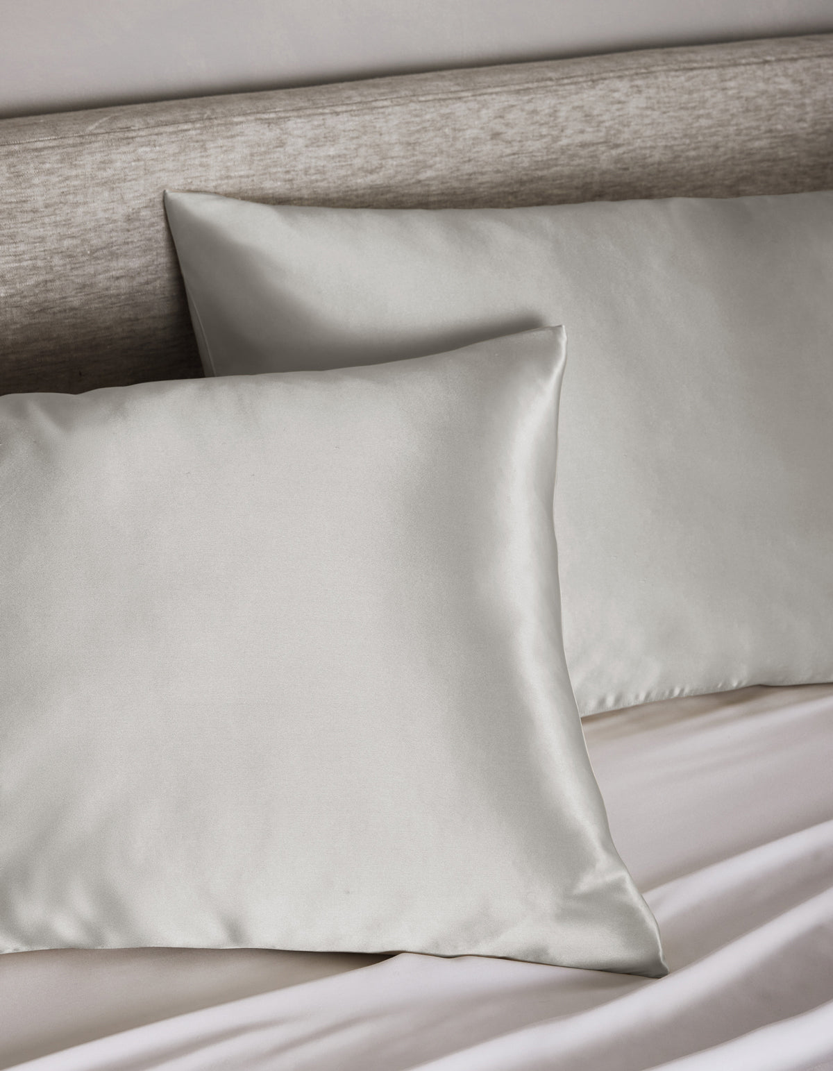 Two Cozy Earth Silk Pillowcases in light gray rest on a matching upholstered headboard and bed, with a neatly tucked white sheet visible below.|Color:Light Grey