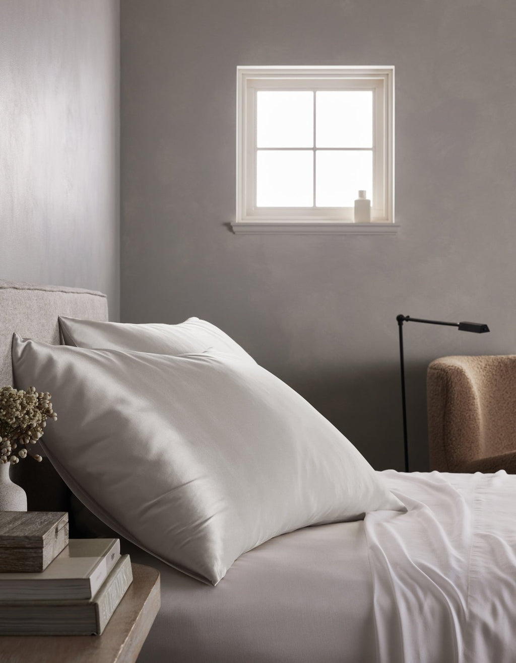 A neatly made bed with Cozy Earth Silk Pillowcases sits beside a stack of books and dried flowers. Soft light streams through a small window, while a beige armchair and black reading lamp are visible in the background.|Color:Light Grey