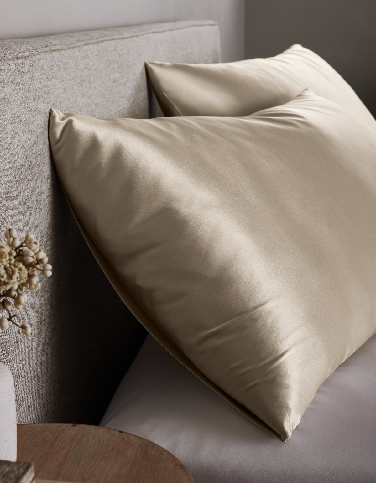 Two large, shiny Cozy Earth Silk Pillowcases in champagne rest against a light upholstered headboard on the bed, with a small round wooden nightstand and a vase of dried flowers nearby.|Color:Oat