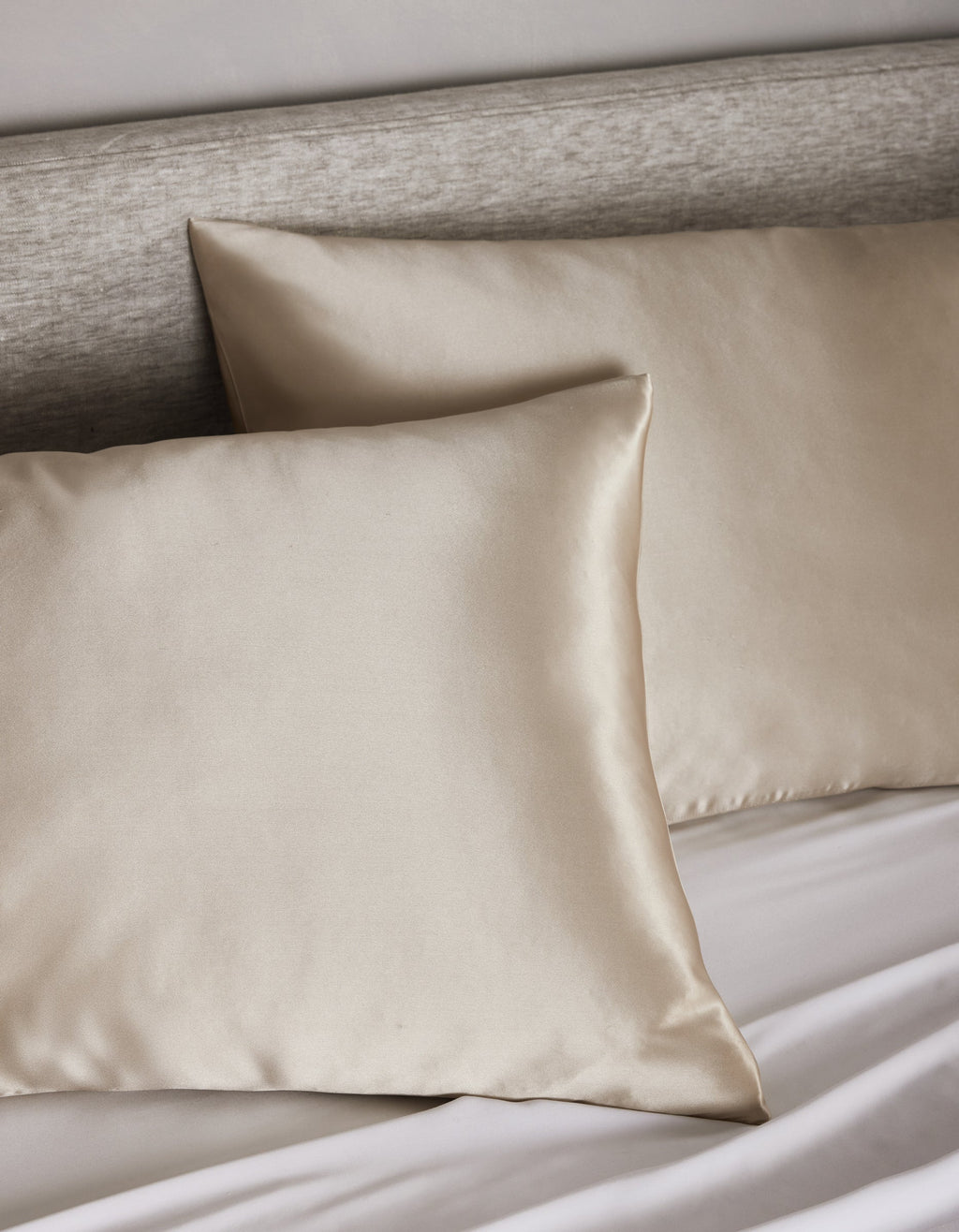 Two Cozy Earth Silk Pillowcases in beige are displayed on a bed with a light gray upholstered headboard and white sheets.|Color:Oat