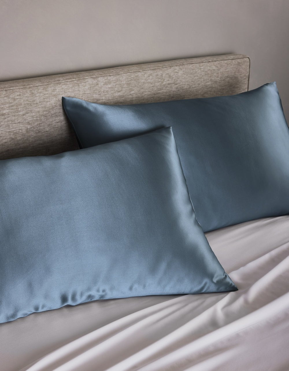 Two Cozy Earth Silk Pillowcases in blue rest on pillows against a light beige upholstered headboard, complemented by light gray sheets. The look is clean and minimalistic.|Color:Pacific Blue