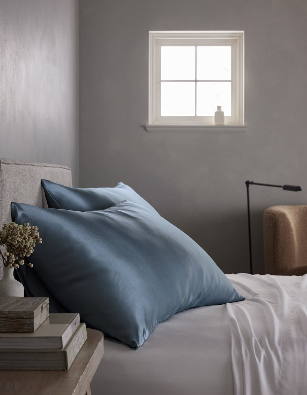A neatly made bed with Cozy Earth Silk Pillowcases and a white sheet sits beside a small window in a softly lit, minimal bedroom. A stack of books and a small vase are arranged on the bedside table.|Color:Pacific Blue