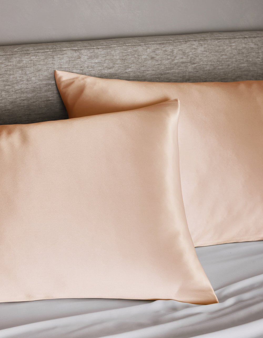 Two Cozy Earth Silk Pillowcases in peach rest on a bed with a light gray upholstered headboard and white sheets.|Color:Peony
