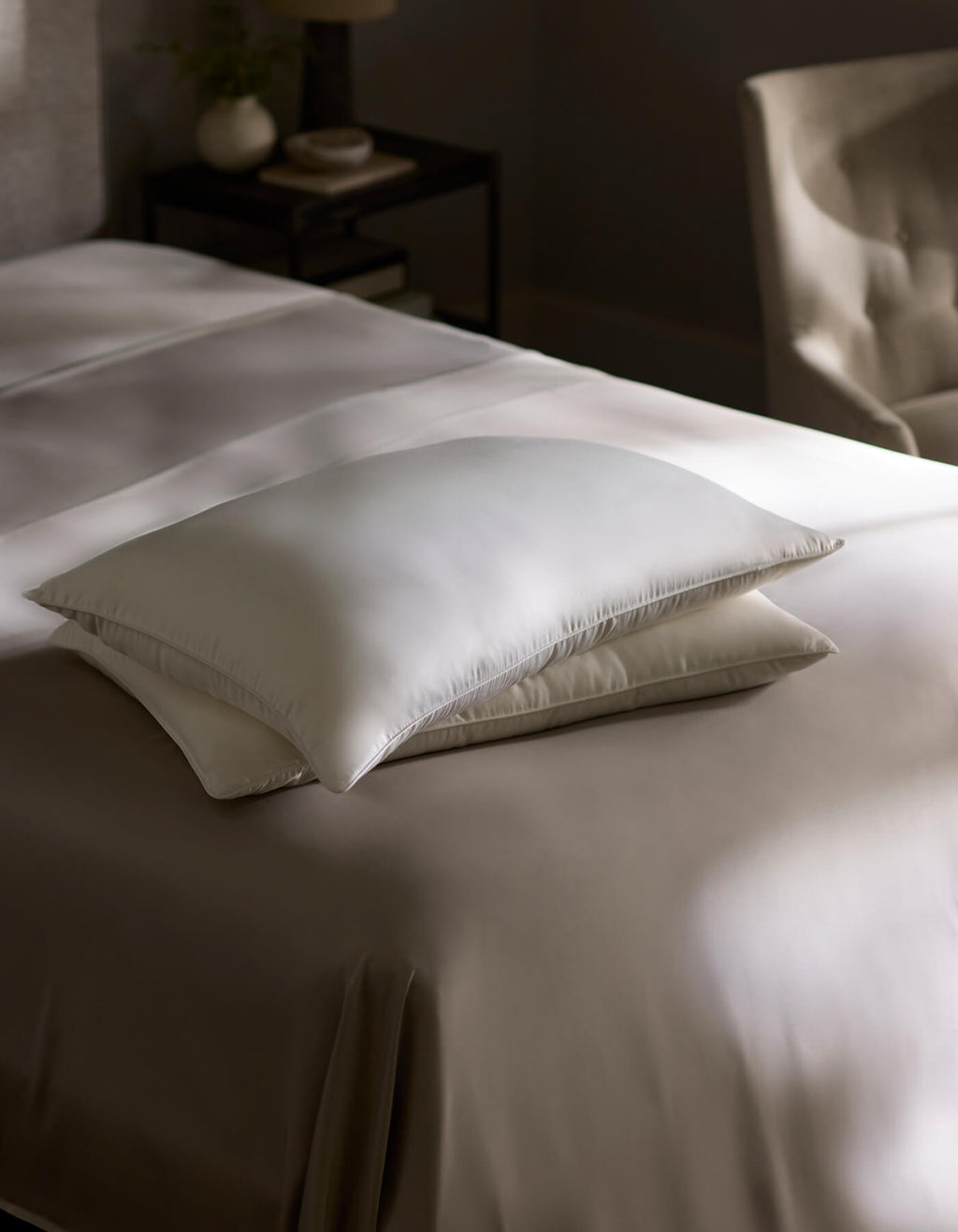 Two Silk Insert Bundle pillows by Cozy Earth are stacked on a neatly made bed with beige sheets in a softly lit, modern bedroom. A nightstand and an upholstered chair appear in the background.|Filling:Extra