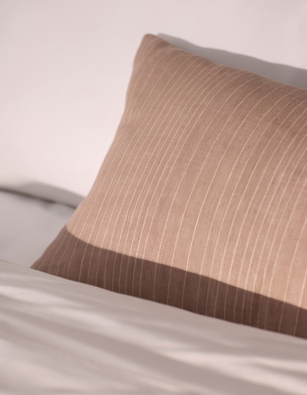 The Cozy Earth Striped Throw Pillow, in beige with subtle vertical stripes, rests against white bedding in soft natural light.|Color:Clay|Size:14" x 30"