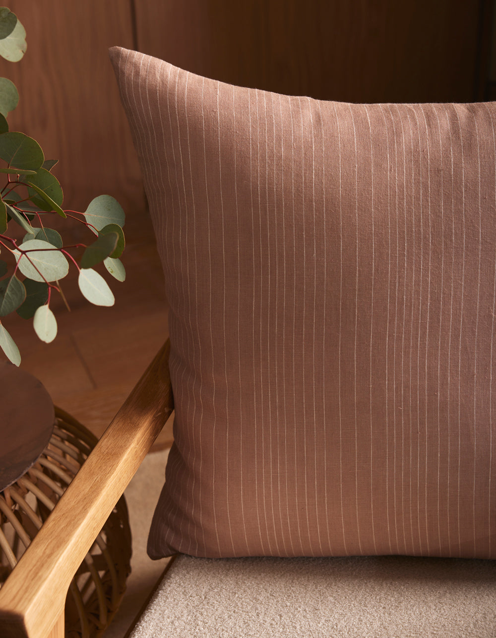 A close-up of the Cozy Earth Striped Throw Pillow in brown on a beige seat, next to a wooden side table with green eucalyptus leaves, creates a warm and cozy indoor atmosphere with soft lighting.|Color:Clay|Size:26" x 26"