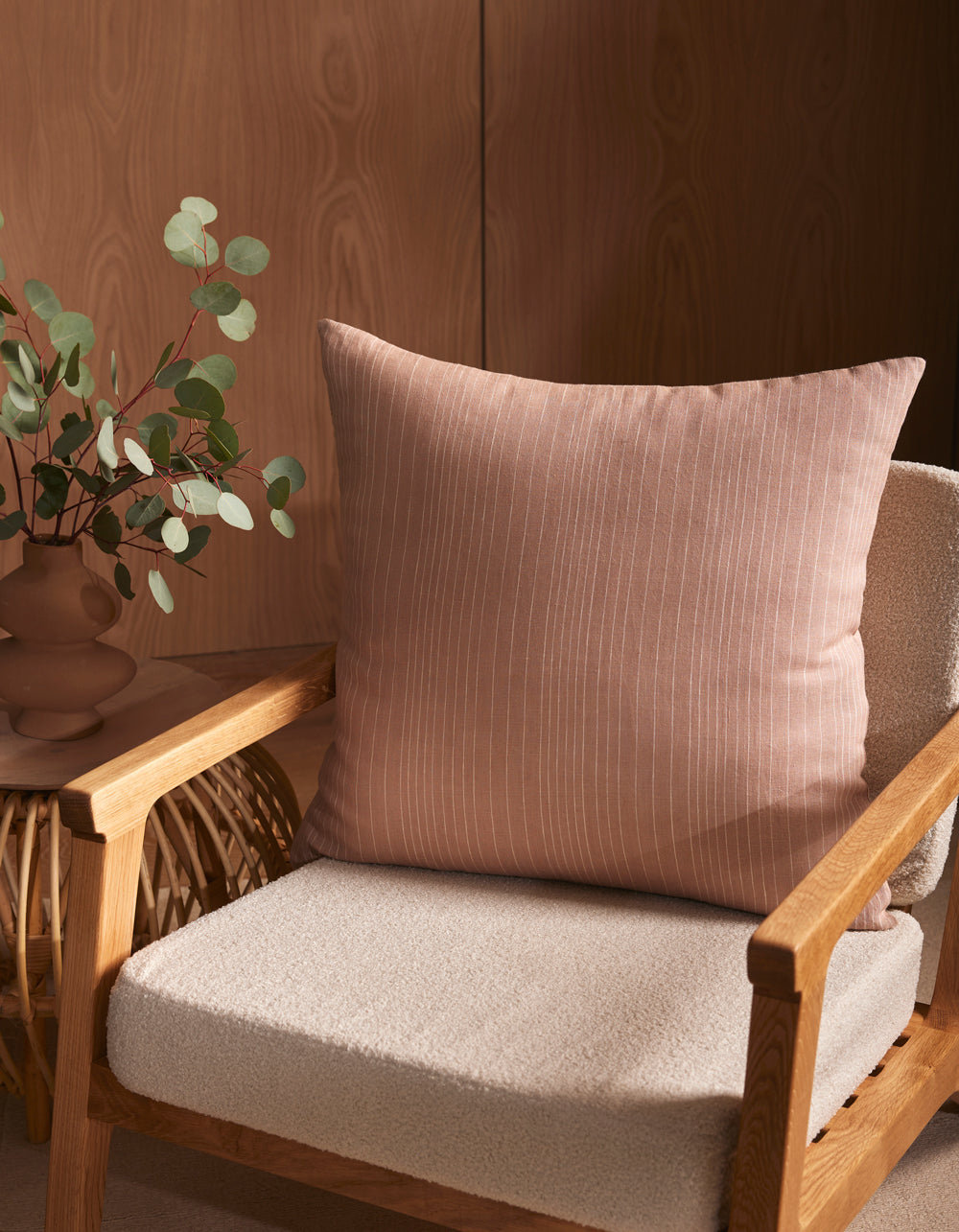 A soft beige armchair with a textured seat cushion features the Striped Throw Pillow by Cozy Earth in pale pink. Beside the chair, a wicker side table holds a vase with eucalyptus branches, all set against a wooden wall.|Color:Clay|Size:20" x 20"