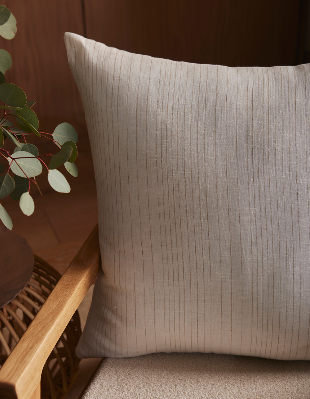 A close-up of the Cozy Earth Striped Throw Pillow on a light cushioned chair, next to a wooden armrest and a round table with eucalyptus leaves in a glass vase.|Color:Ecru|Size:26" x 26"