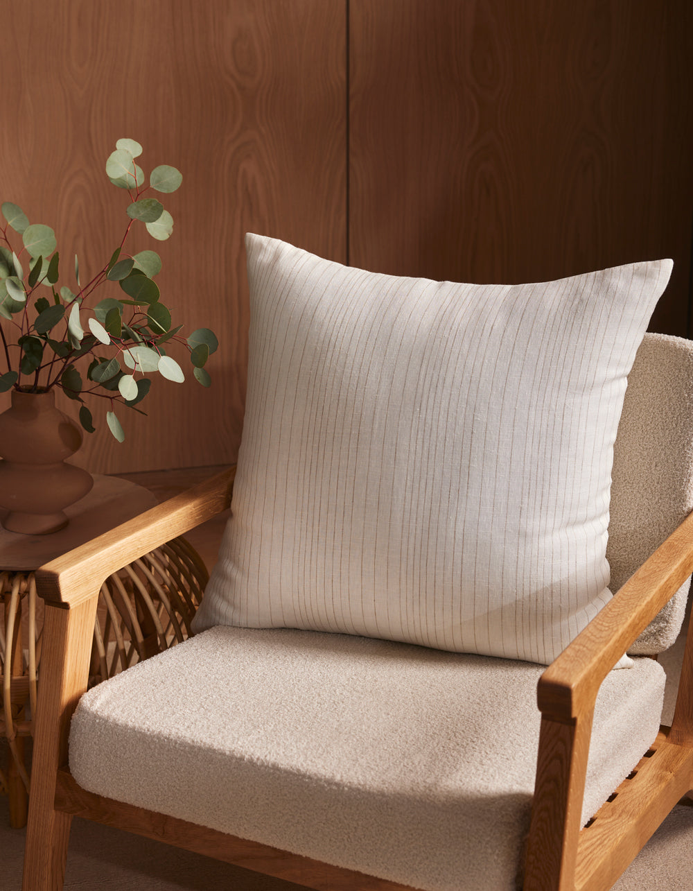 A Cozy Earth Striped Throw Pillow rests on a wooden armchair with a textured cream seat. Beside it, a small woven side table displays a brown vase filled with green eucalyptus branches.|Color:Ecru|Size:26" x 26"