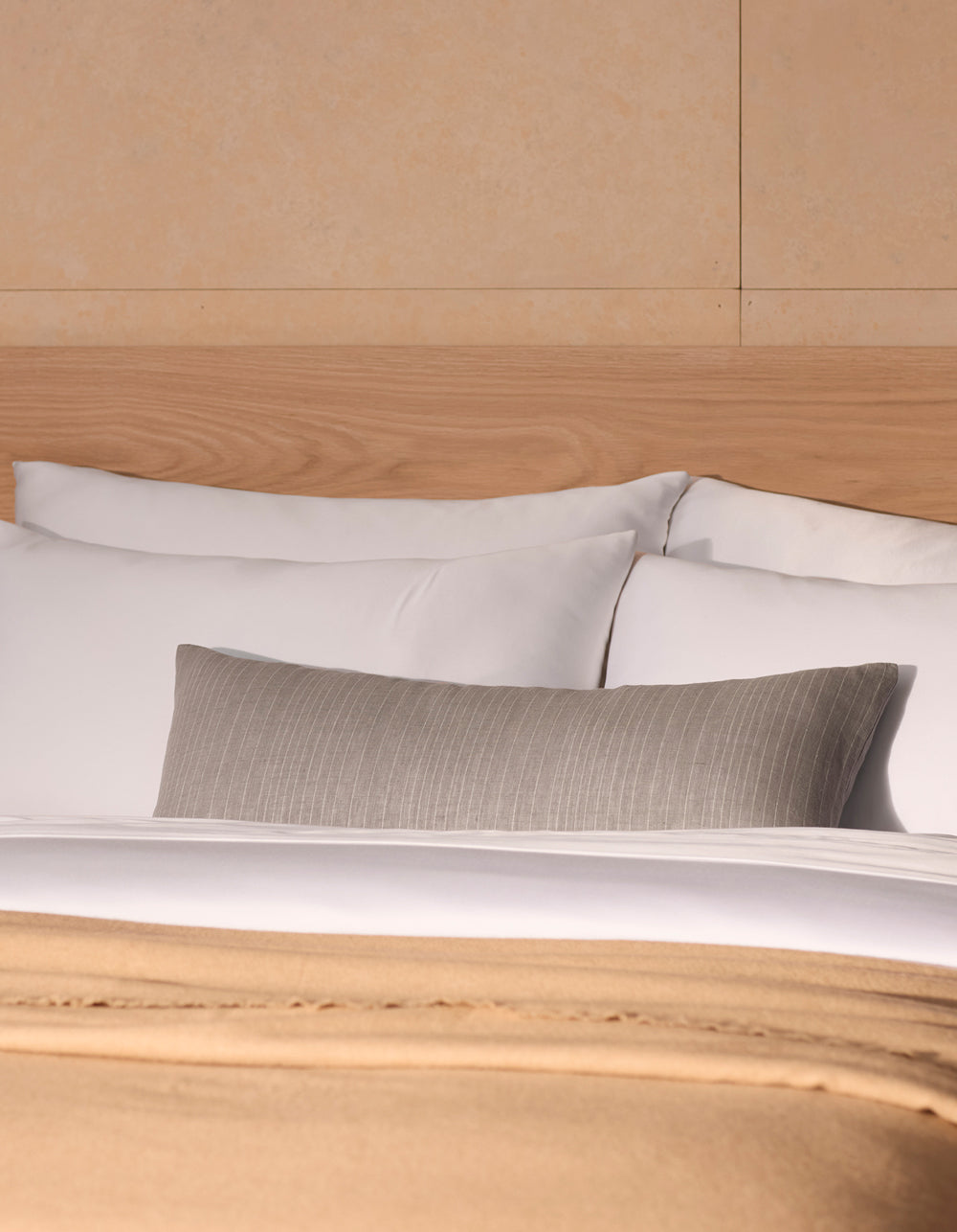 A neatly made bed features Cozy Earth’s Striped Throw Pillow with white pillows, a beige blanket, and a gray cushion, set against a light wood headboard and cream-colored wall.|Color:Grey|Size:14" x 30"