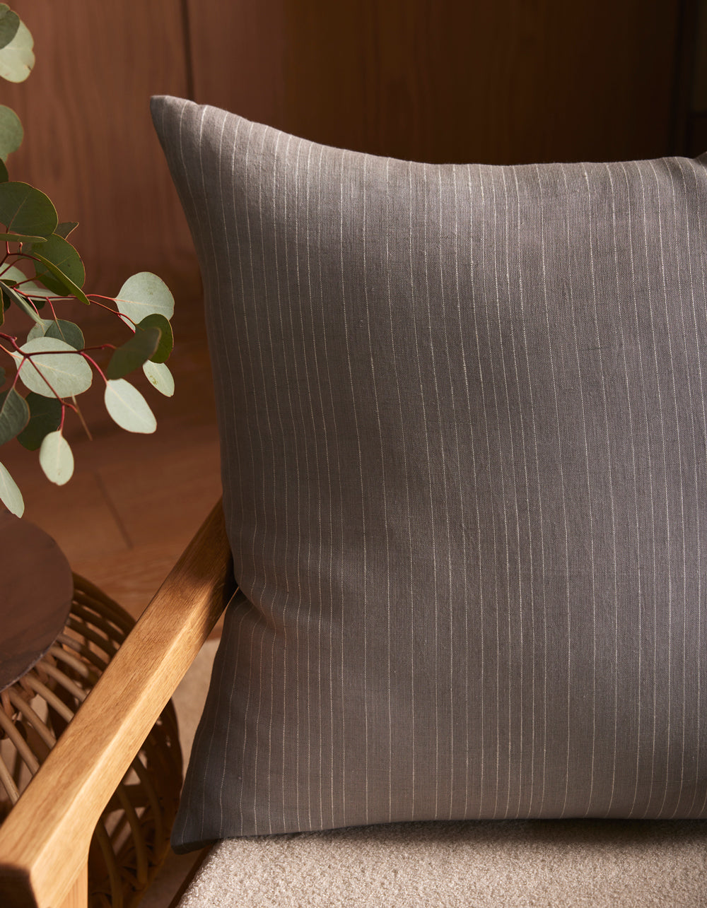 The Striped Throw Pillow by Cozy Earth, featuring thin white pinstripes on gray, sits on a light beige seat. A wooden chair and green plant are nearby, while warm indoor lighting adds to the cozy atmosphere.|Color:Grey|Size:24" x 24"
