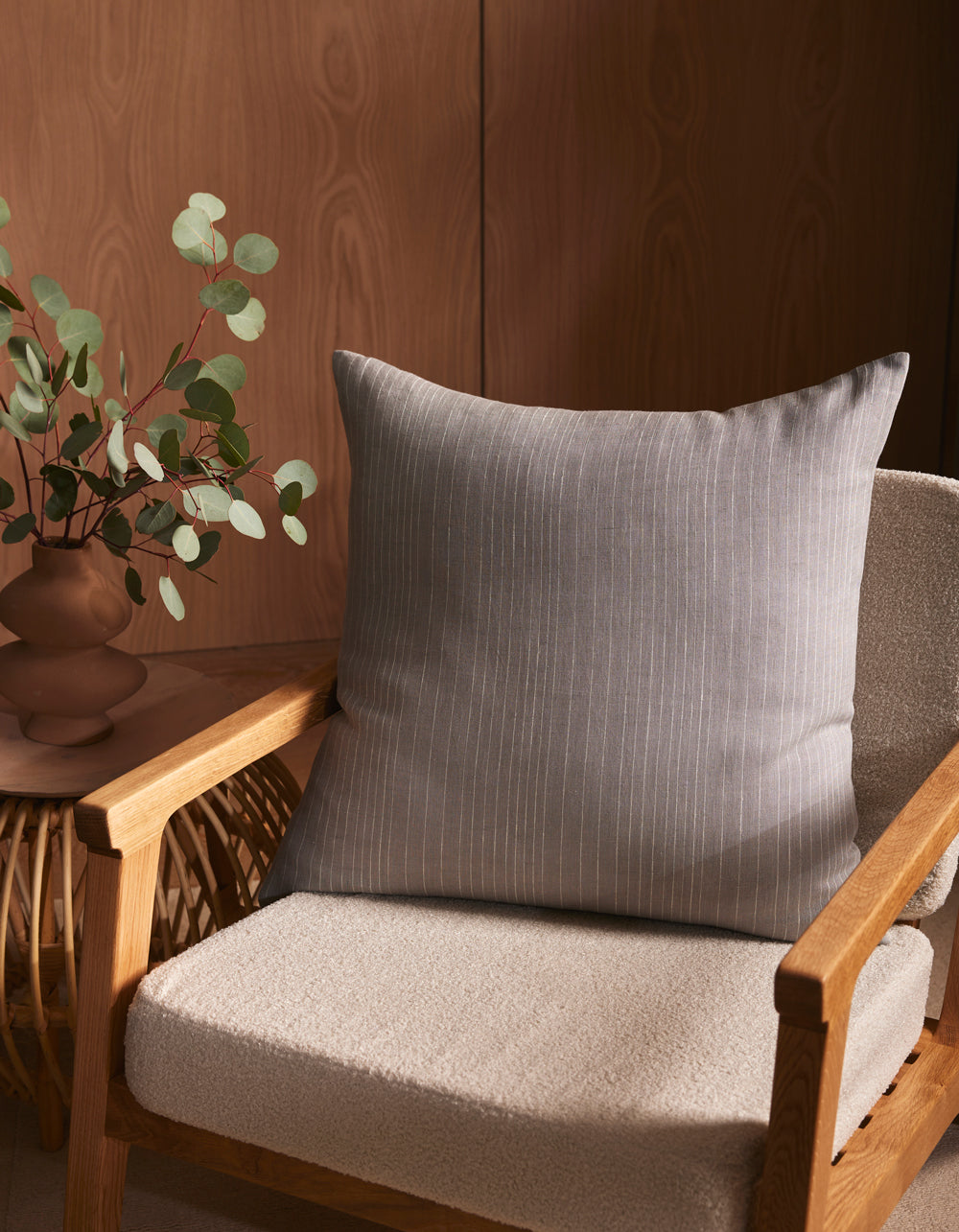 A wooden armchair with a light beige cushion features the Striped Throw Pillow by Cozy Earth and sits by a small table with a brown vase of eucalyptus, set against a wooden wall.|Color:Grey|Size:26" x 26"