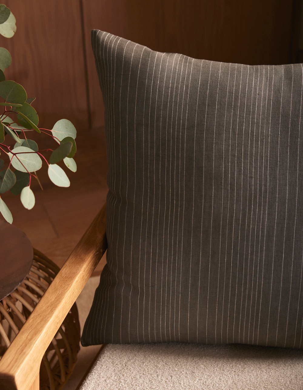 A Cozy Earth Striped Throw Pillow in dark gray with thin white stripes sits on a beige cushioned wooden chair beside a round table decorated with green eucalyptus leaves.|Color:Smoke|Size:24" x 24"