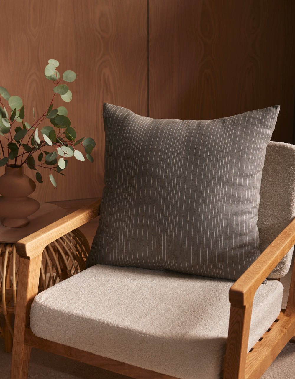 A wooden armchair with a textured, light beige cushion and the Cozy Earth Striped Throw Pillow sits next to a small table with a brown vase of eucalyptus branches, all set against a wooden wall.|Color:Smoke|Size:26" x 26"