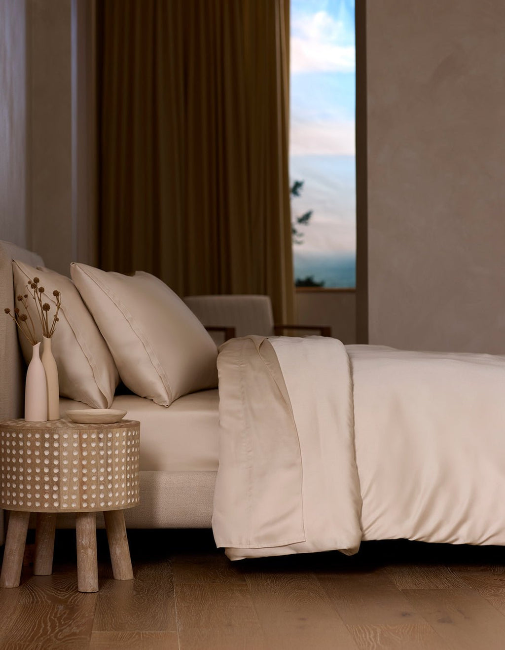 A neatly made bed with the Cozy Earth Bamboo Sheet Set in cream sits next to a round wooden nightstand holding two vases of dried flowers, as soft natural light streams through a window with beige curtains.|Color:Creme