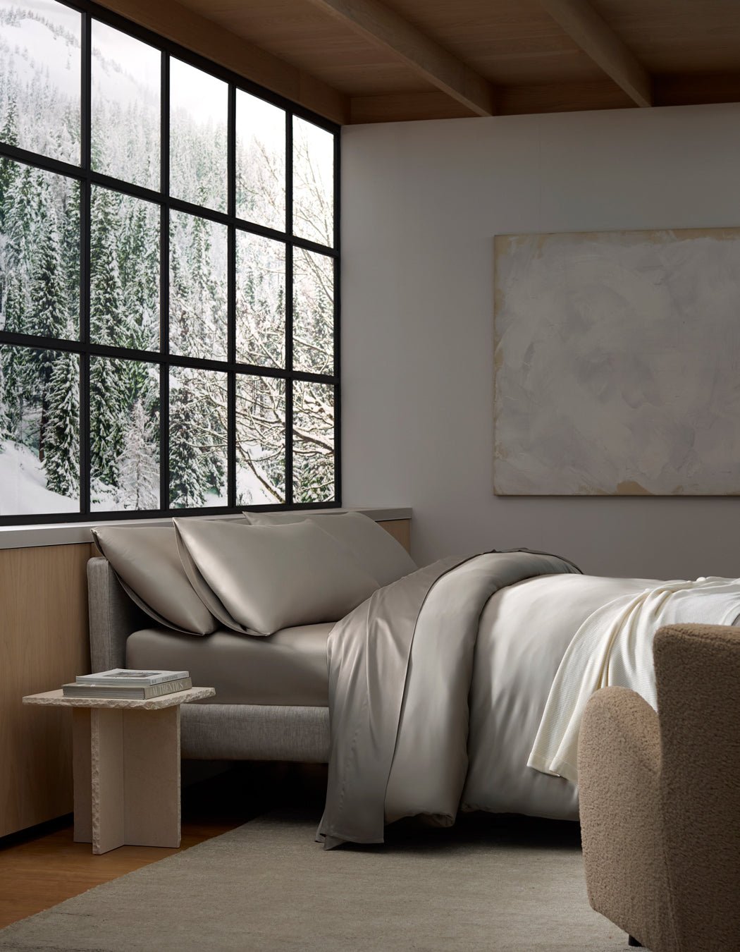 A cozy bedroom with a large window framing snowy pine trees outside. The bed features Cozy Earth's Bamboo Bedding Ultimate Bundle in gray, and there’s a beige chair and side table with books, creating a calm, minimalist retreat.|Color:Dove Grey