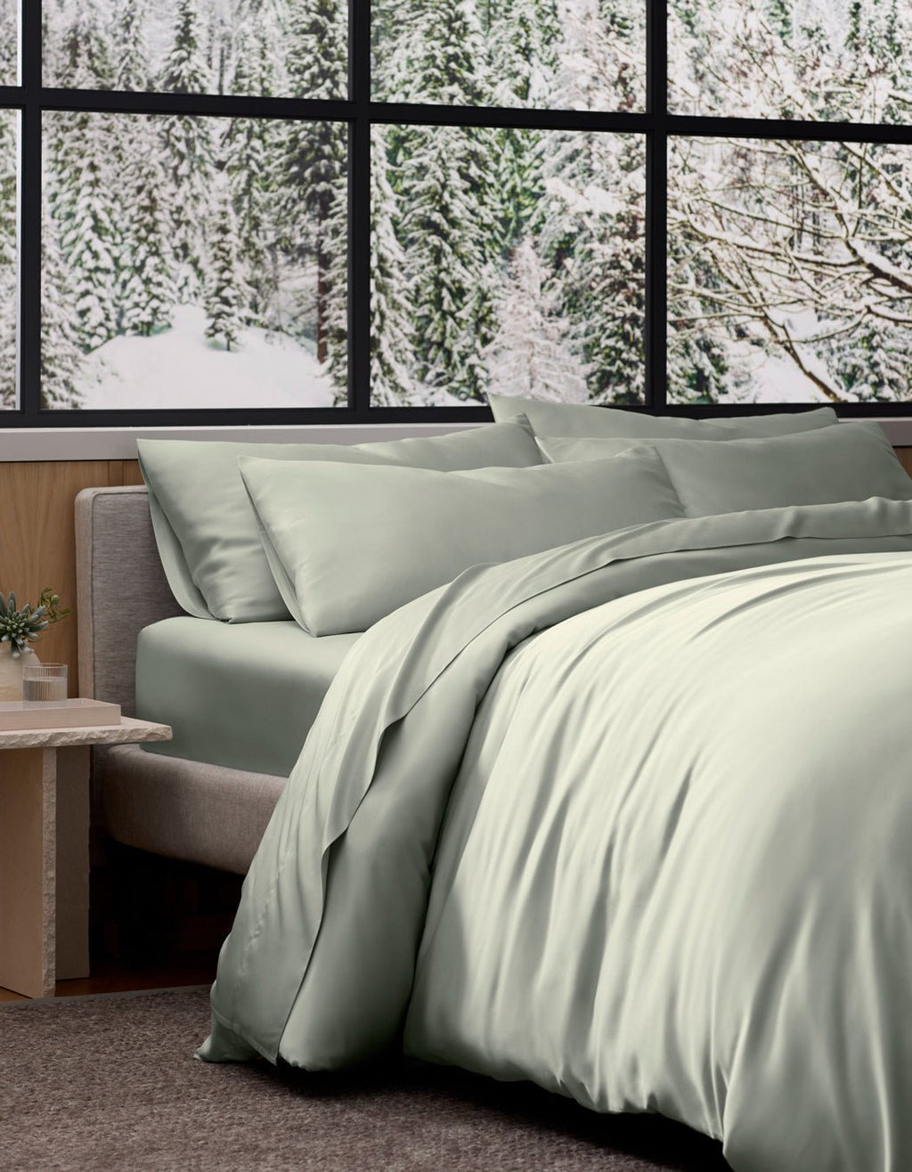 A neatly made bed with a light sage green Bamboo Duvet Cover by Cozy Earth sits by a window overlooking a snowy pine forest, with a small potted plant on the side table.|Color:Laurel