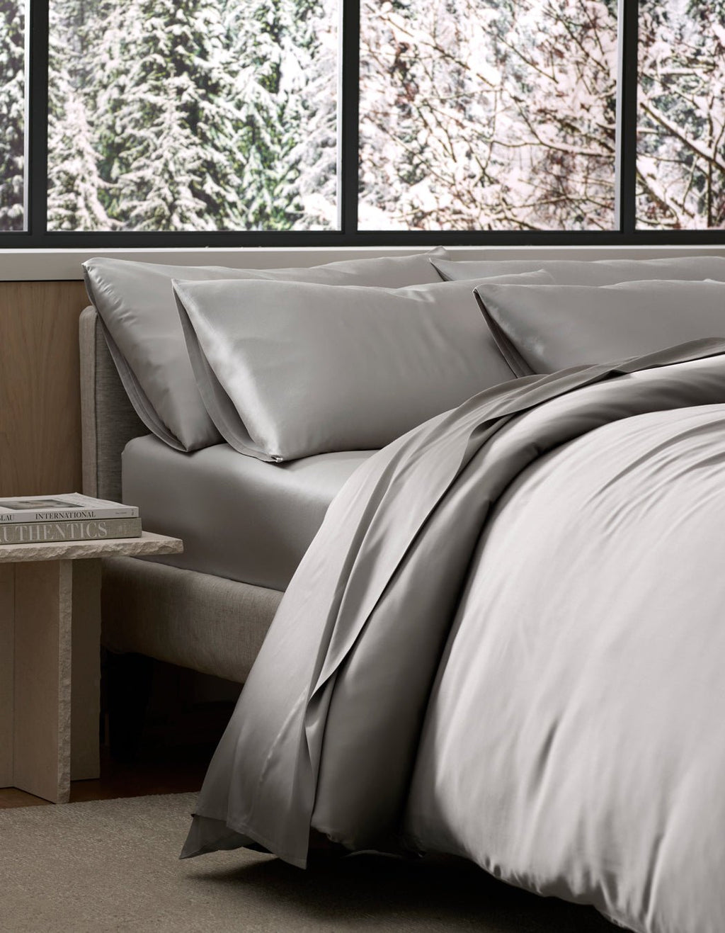 A bed dressed in Cozy Earth’s Bamboo Bedding Ultimate Bundle in light gray sits by a window with a snowy forest view, while a small bedside table holds a stack of books.|Color:Light Grey