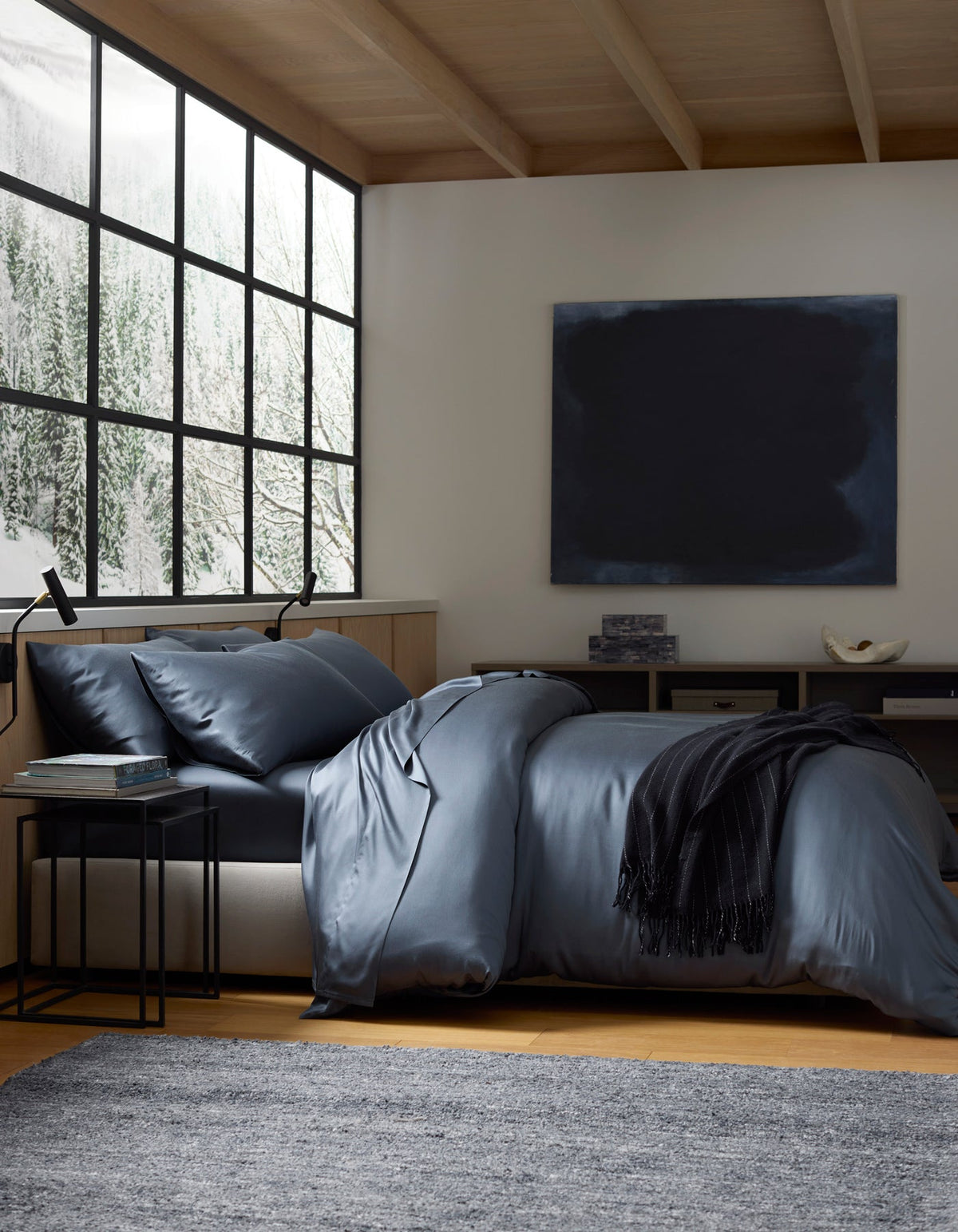 A modern bedroom with a large window showing snowy trees, a bed made with Cozy Earth's Bamboo Bedding Deluxe Bundle in dark blue, a black throw, side table, grey rug, and a dark abstract painting on the wall.|Color:Pacific Blue