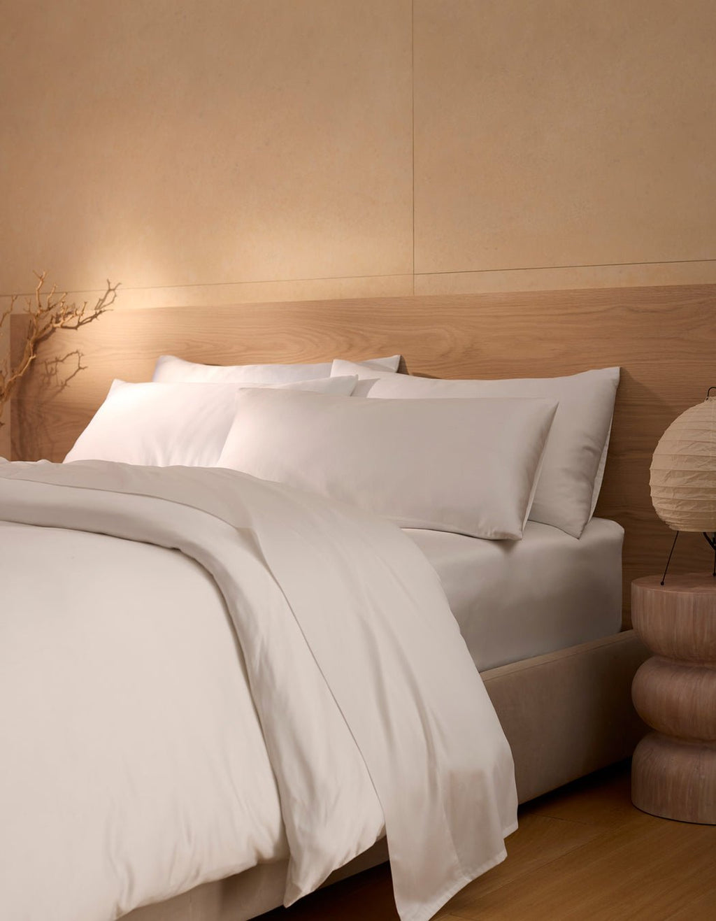 A minimalist bedroom featuring the Cozy Earth Bamboo Bedding Ultimate Bundle—white sheets, pillows, and comforter—next to a wooden nightstand with a round lamp and decorative branch in soft lighting.|Color:White