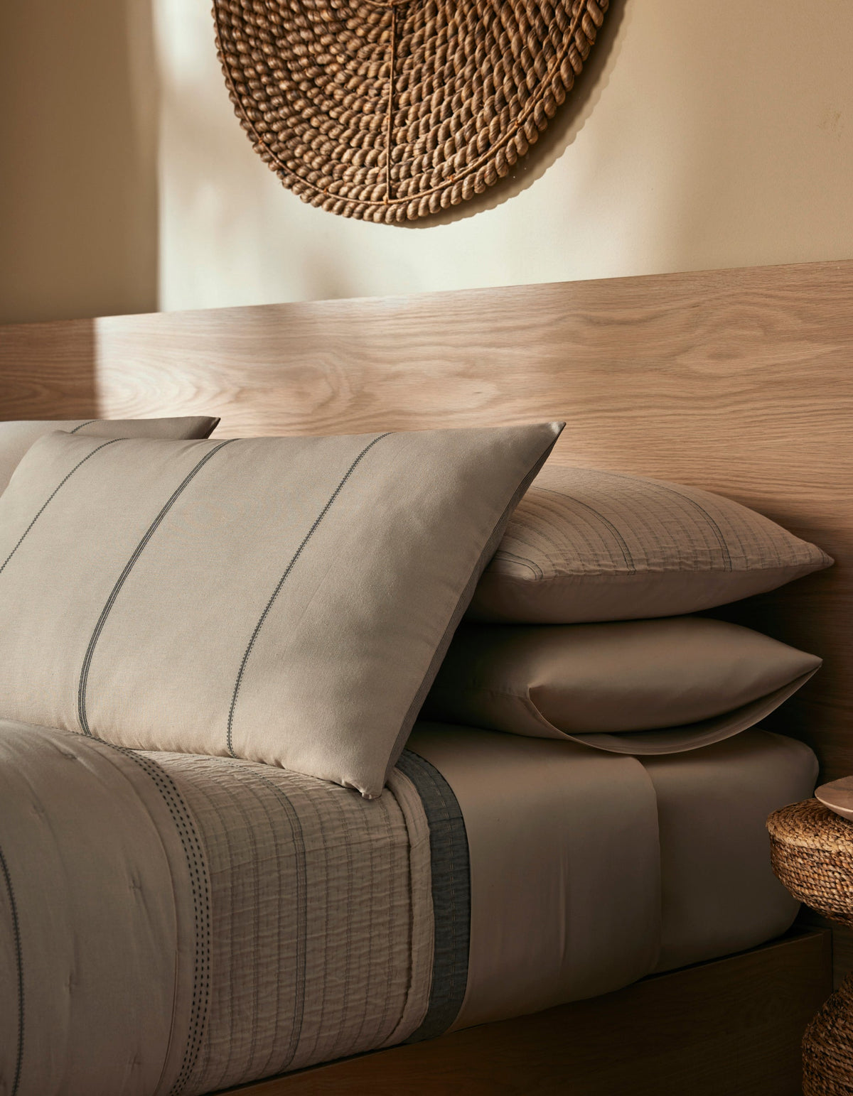 A cozy bedroom featuring the Cozy Earth Baja Double Sided Sham with taupe and gray bedding, stacked pillows, a light wood headboard, and a woven wall piece above, all softly lit for a warm ambiance.|Color:Coal Stripe/Striped Alabaster