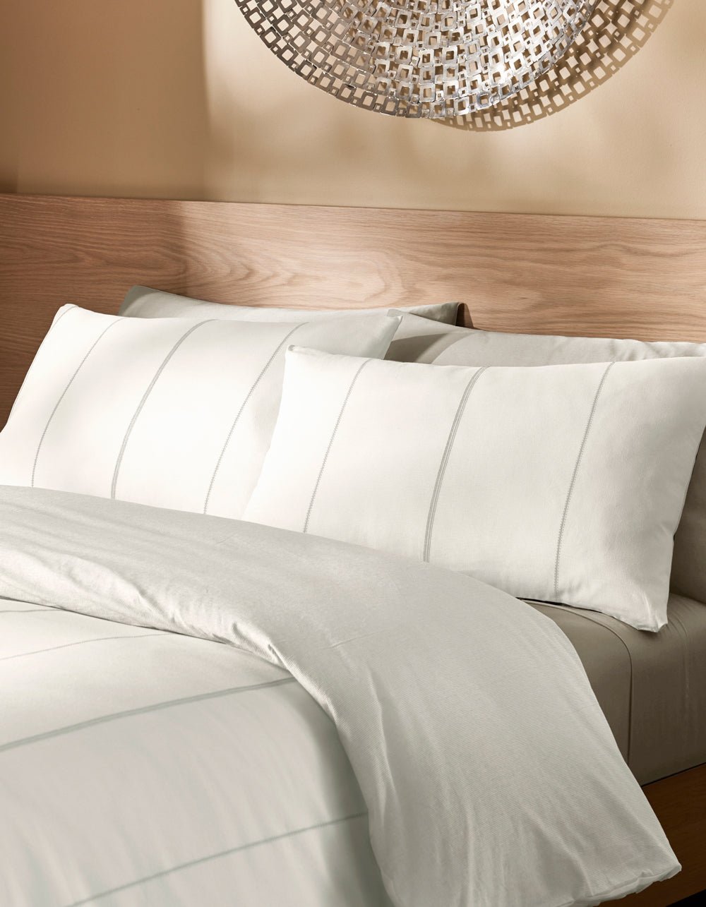 A bed featuring the Cozy Earth Baja Duvet Cover in white, with subtle stripe patterns on the bedding and pillows, is set against a light wooden headboard and beige wall adorned with metallic wall art.|Color:Laurel Stripe/Striped Bone