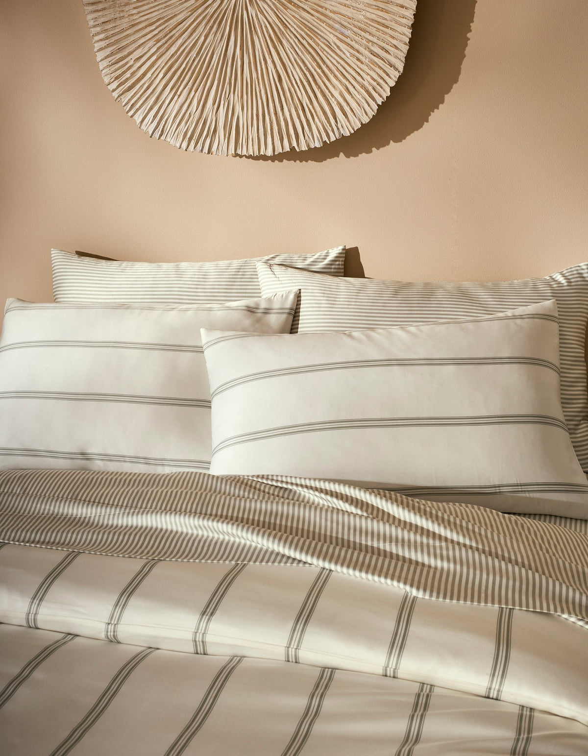A neatly made bed features Cozy Earth Bamboo Pillowcases in cream and light gray stripes, accented by matching bedding and set against a beige wall with a textured, fan-shaped decoration above the headboard.|Color:Baja Stripe in Laurel