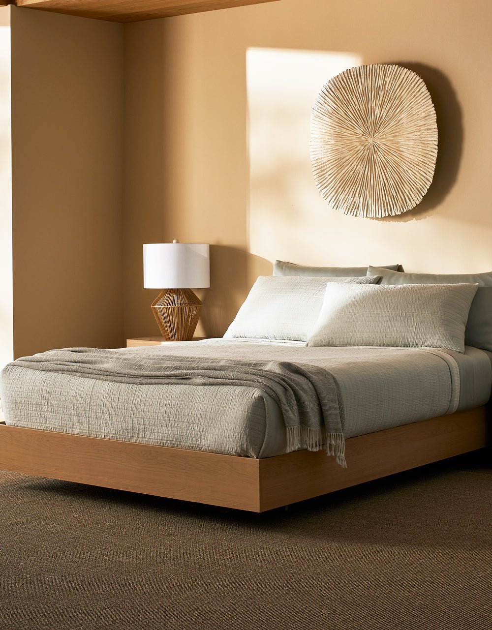 A modern bedroom featuring the Cozy Earth Baja Matelasse Bundle on a low wood bed, pale gray bedding with a gray throw, a wooden nightstand with lamp, and a decorative wall piece. The space showcases neutral, earthy tones.|Color:Bone/Laurel