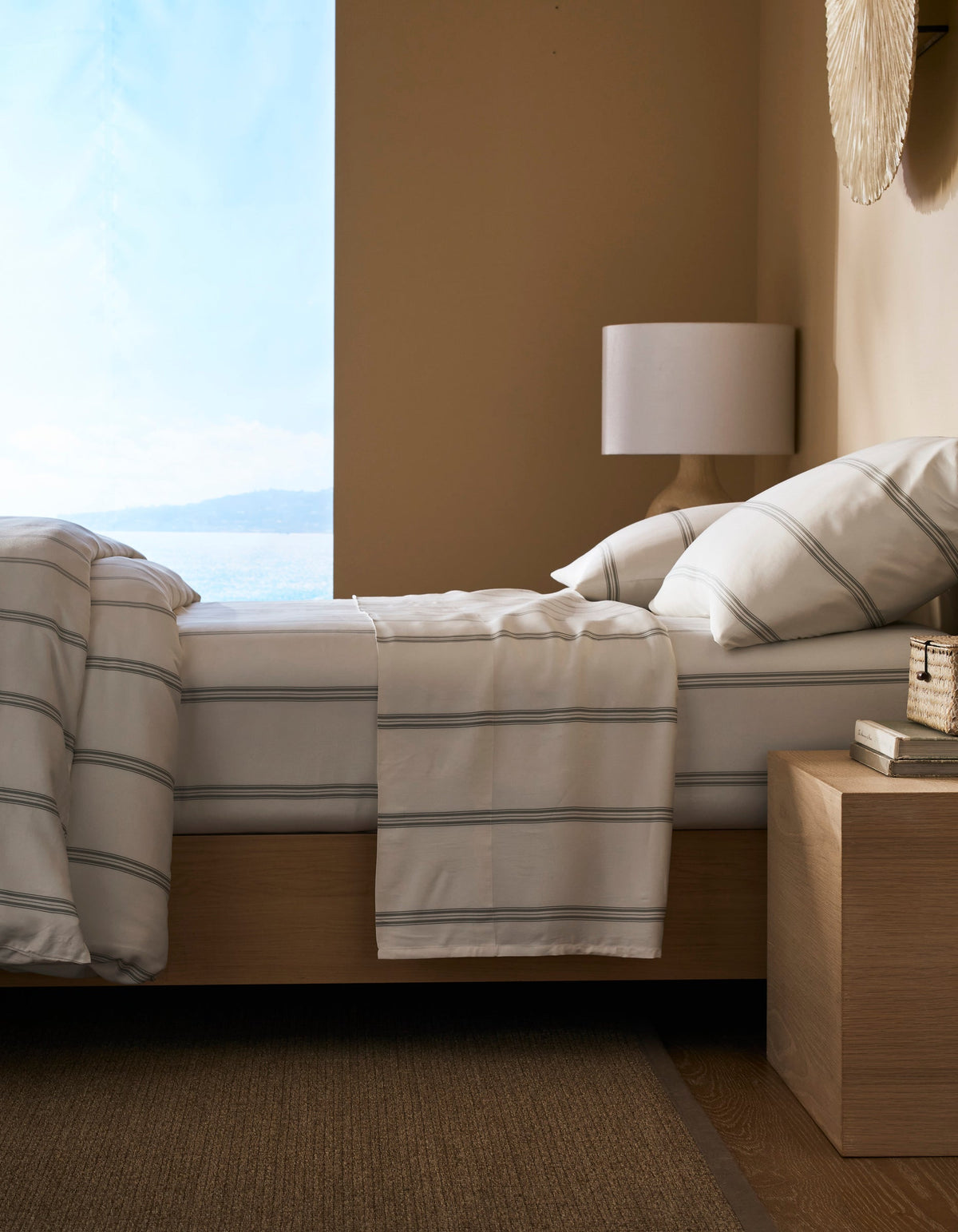 A modern bedroom featuring a neatly made bed with Cozy Earth’s Bamboo Sheet Set, a wooden nightstand with books and decor, a white lamp, and a large window offering views of water and sky.|Color:Baja Stripe in Laurel
