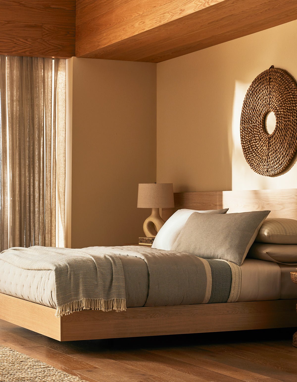 A modern bedroom features Cozy Earth's Baja Quilt Bundle on a wooden platform bed, with neutral tones, a woven wall hanging, a beige lamp on a nightstand, and sunlight filtering through sheer curtains onto wood floors.|Color:Coal Stripe/Striped Alabaster