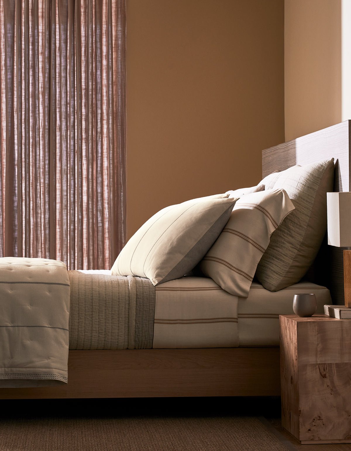 A neatly made bed with the Cozy Earth Baja Matelasse Coverlet stands beside a wooden nightstand holding a lamp and cup, in a softly lit room with sheer curtains and warm-toned walls.|Color:Ebony/Taupe