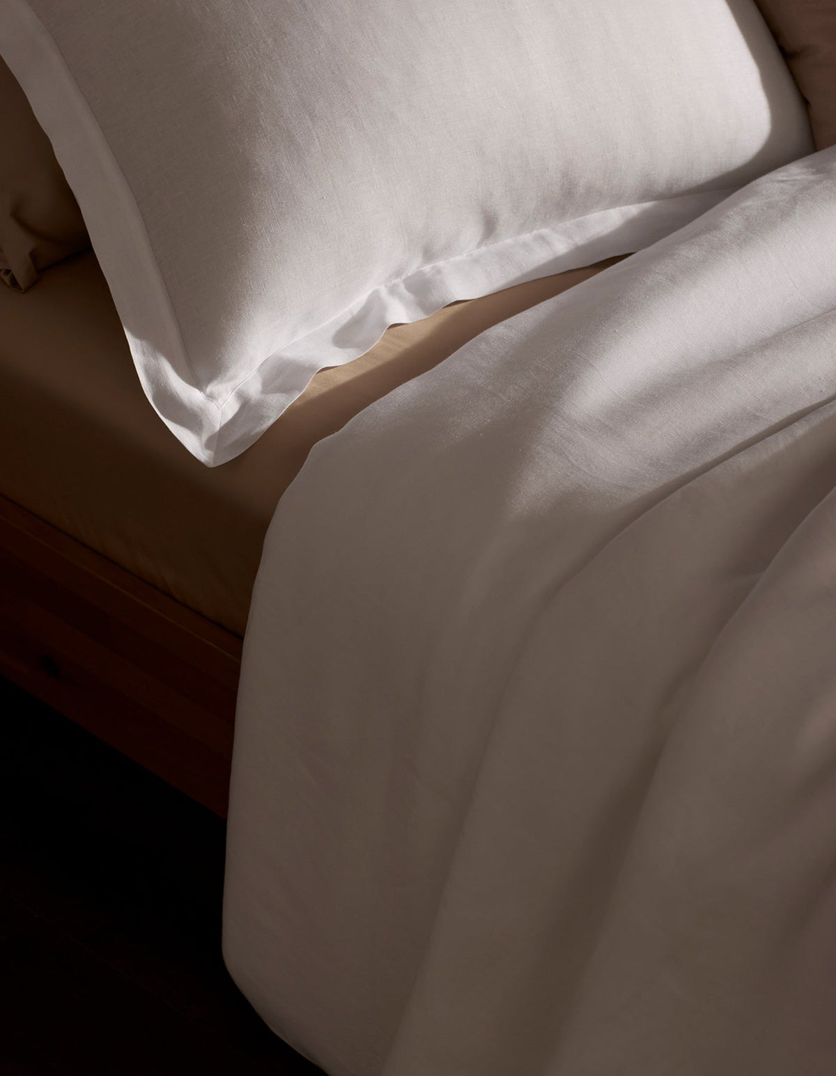 A neatly made bed features the Cozy Earth Linen Duvet Set—a white pillow and sheet atop a beige fitted sheet—all bathed in soft natural light.|Color:White