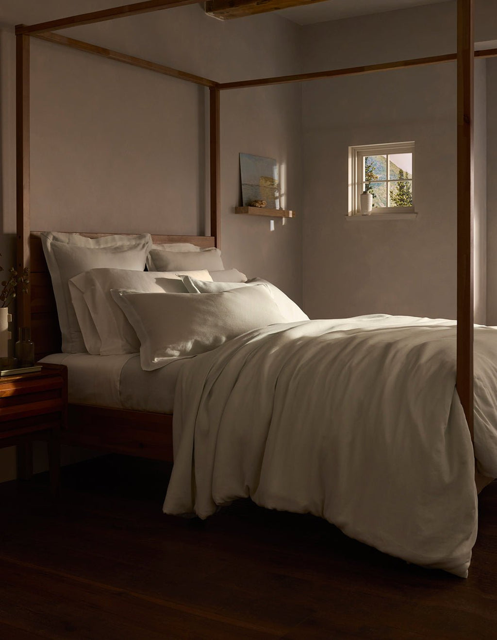 A cozy bedroom features a wooden four-poster bed with layered white bedding, including the Linen Duvet Cover by Cozy Earth. Warm light from a small window softly illuminates the neutral tones, creating an inviting atmosphere.|Color:Mist