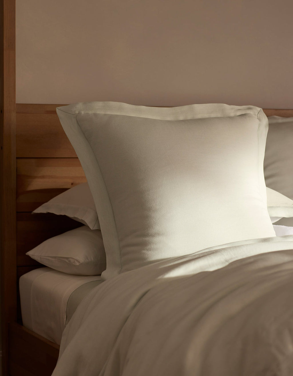 A bed featuring Cozy Earth Linen Shams is neatly made, with a large pillow propped against a wooden headboard and bathed in soft, natural light.|Color:Mist