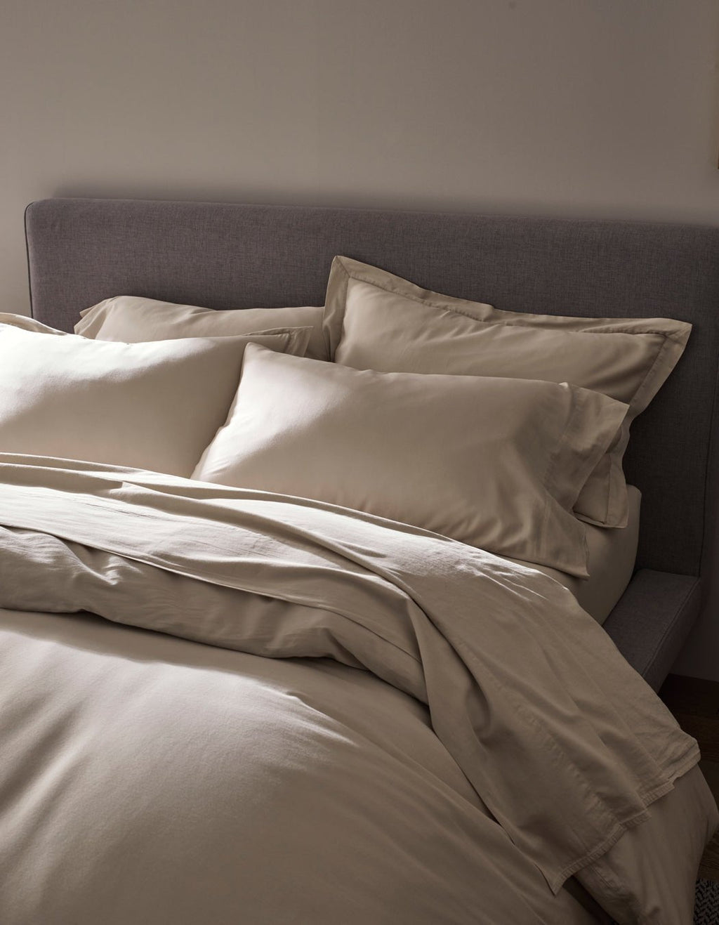 A neatly made bed features the Cozy Earth Soft-Wash Cotton Duvet Cover in cream, set against a grey upholstered headboard and softly lit for a cozy, minimalistic bedroom ambience.|Color:Driftwood