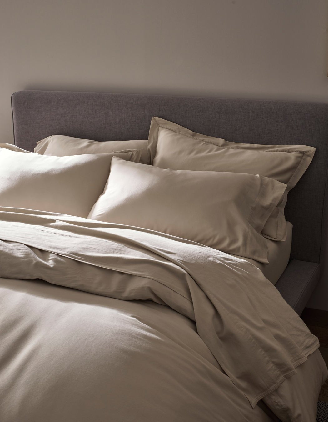 A neatly made bed features the Cozy Earth Soft-Wash Cotton Duvet Cover in cream, set against a grey upholstered headboard and softly lit for a cozy, minimalistic bedroom ambience.|Color:Driftwood