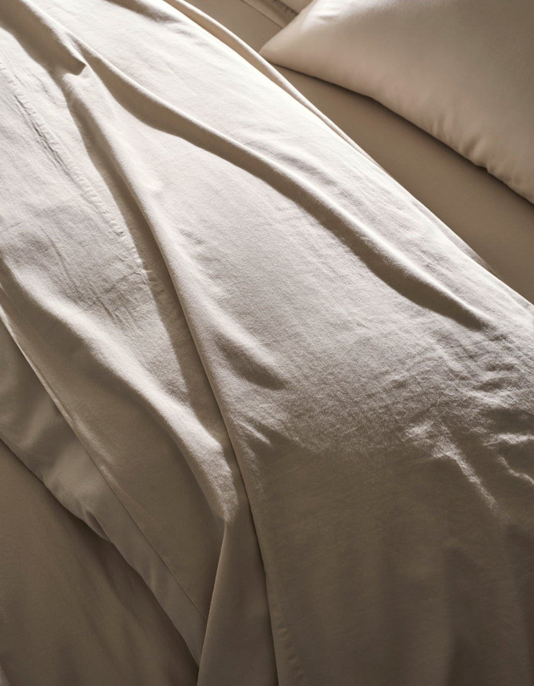 Close-up of a neatly made bed with Cozy Earth's Soft-Wash Cotton Duvet Cover in beige, featuring a matching pillow. The soft, smooth fabric is highlighted by gentle natural light, showcasing its luxurious texture.|Color:Driftwood