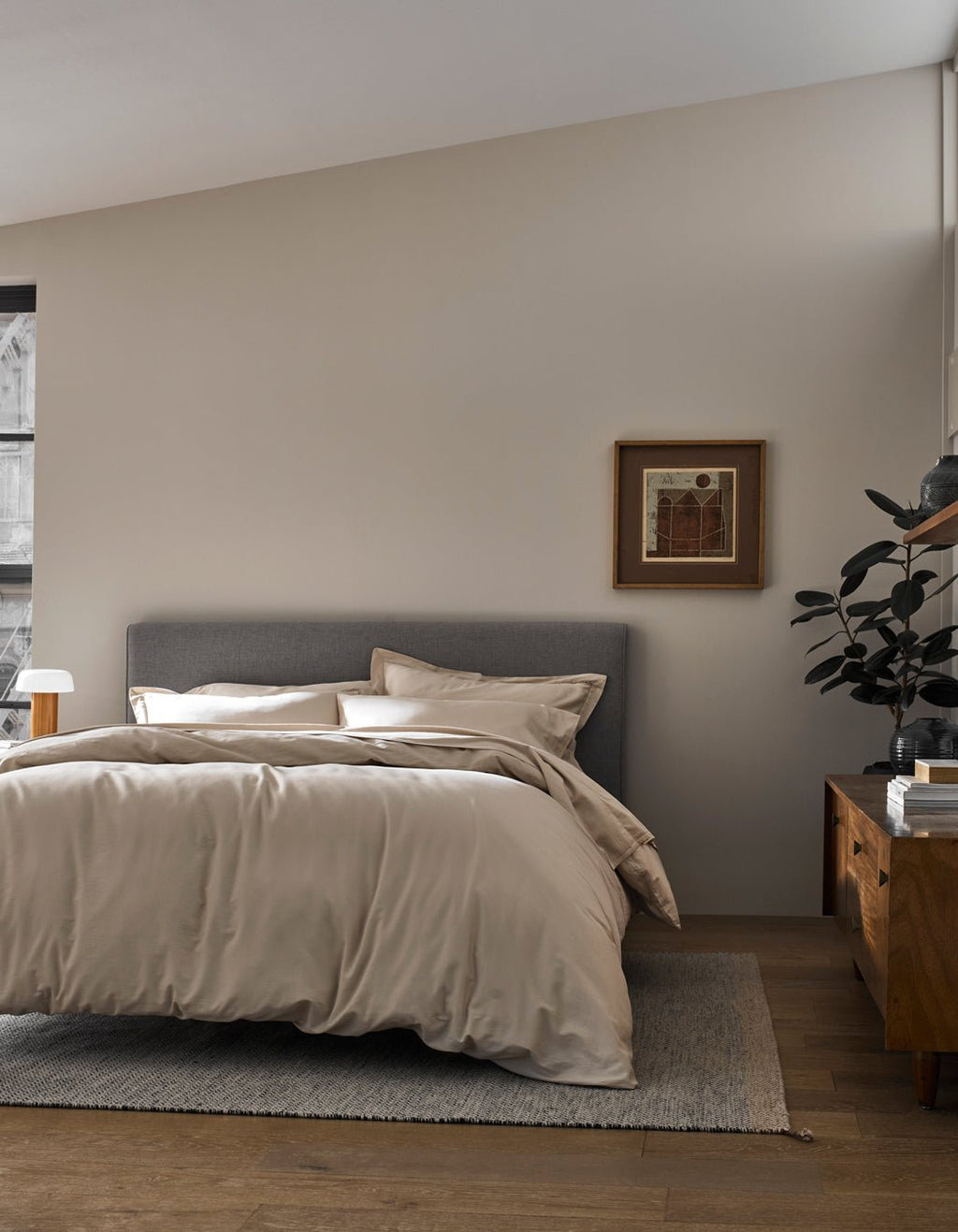 A modern bedroom features a gray upholstered bed with Cozy Earth's Soft-Wash Cotton Duvet Cover in beige, a wooden nightstand holding a plant and lamp, a framed picture on a light-colored wall, all atop wood flooring with a neutral rug.|Color:Driftwood