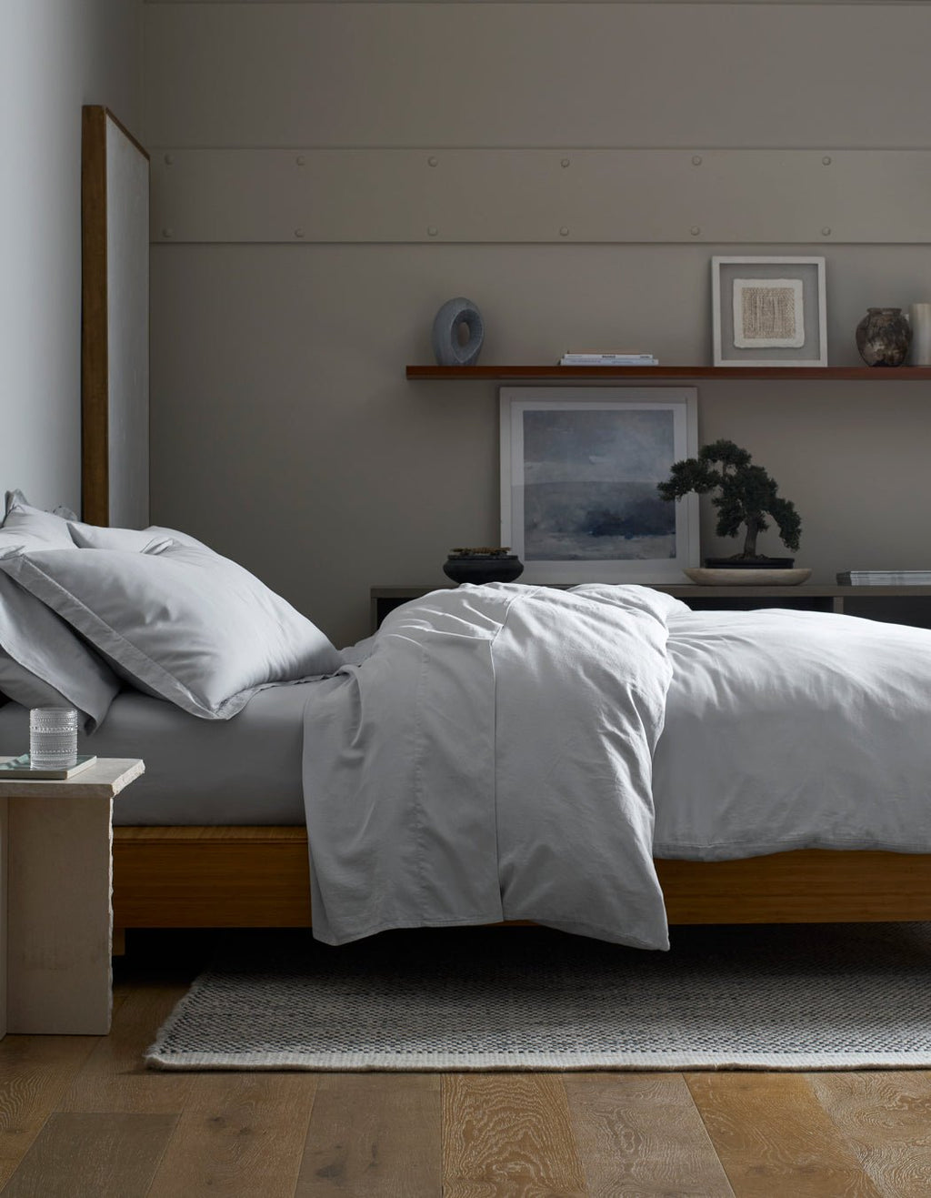 A minimalist bedroom features a neatly made bed with a Cozy Earth Soft-Wash Cotton Duvet Cover in light gray on a wooden frame. Above a dark dresser, art, decor, and a small bonsai are arranged on a floating shelf.|Color:Shore
