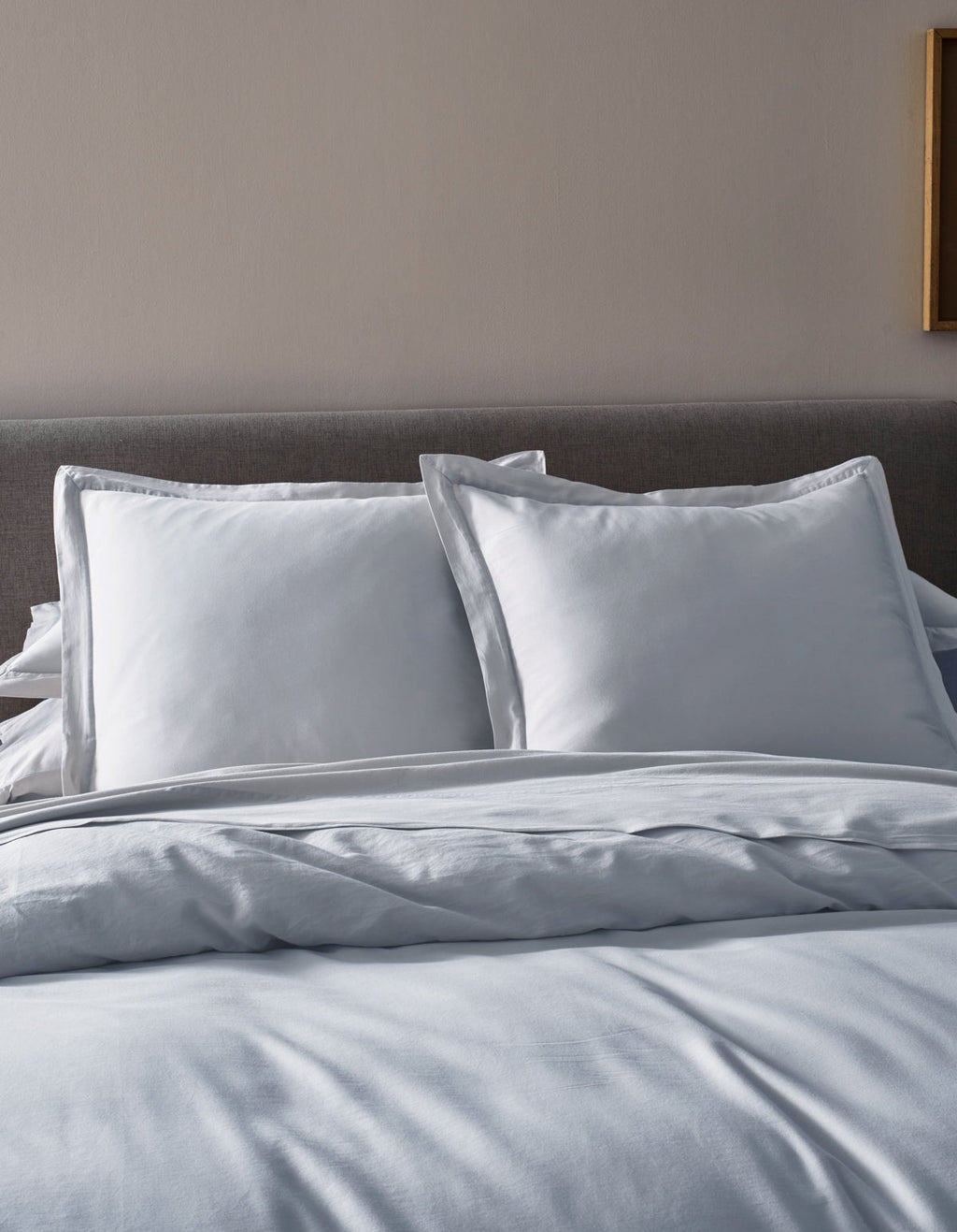 A neatly made bed features Cozy Earth’s Soft-Wash Cotton Duvet Cover, a gray upholstered headboard, two large white pillows, and white sheets in a softly lit bedroom.|Color:Shore