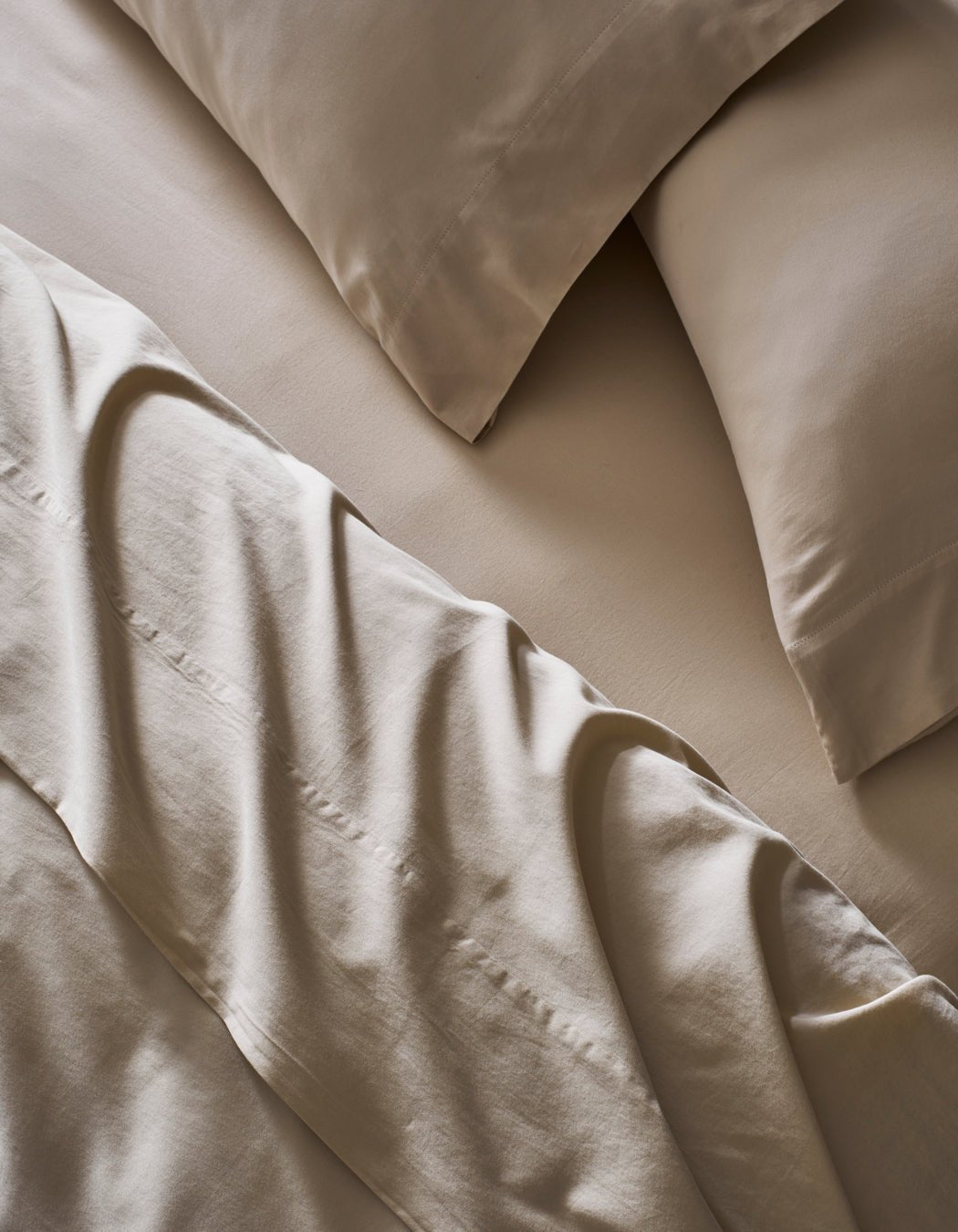 A close-up of a neatly made bed features Cozy Earth's Soft-Wash Cotton Sheet Set in beige. The slightly rumpled sheets and pillowcases enhance the soft, textured look of the smooth fabric.|Color:Driftwood