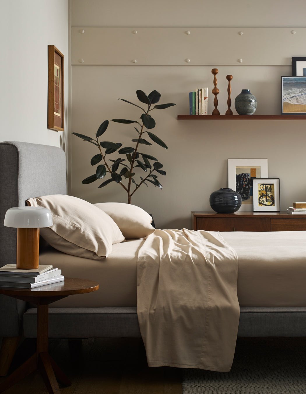 A neatly made bed with Cozy Earth Soft-Wash Cotton Pillowcases sits beside a wooden nightstand with a white lamp. A large green plant, artwork, and decor items decorate the dresser and a wooden shelf on the wall.|Color:Driftwood