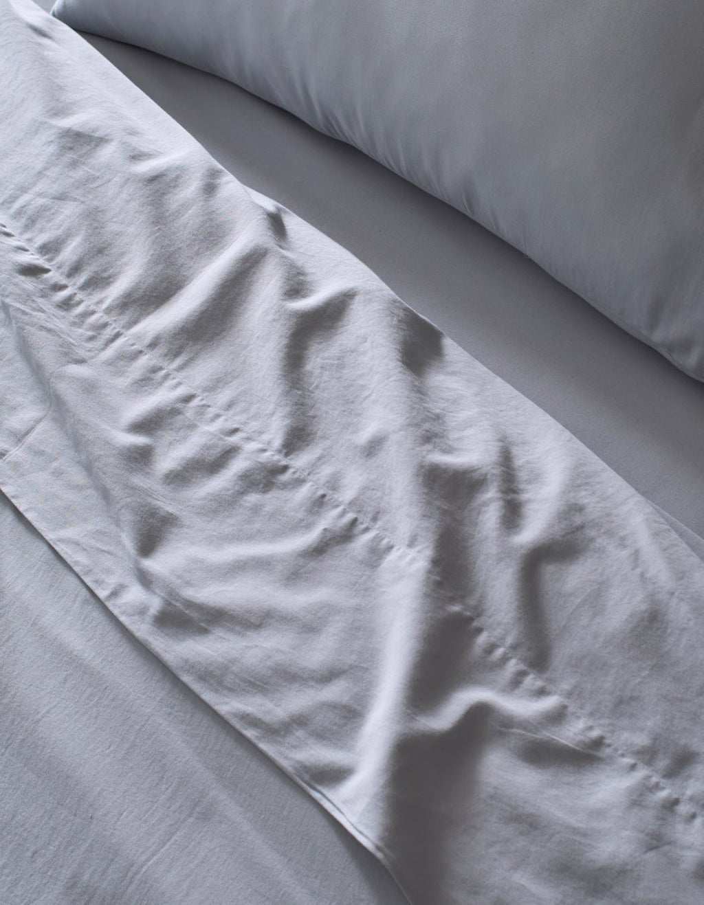 Close-up of a neatly made bed featuring Cozy Earth's Soft-Wash Cotton Sheet Set in light grey. Slight wrinkles add texture to the soft, clean fabric, giving the bedding an inviting look.|Color:Shore